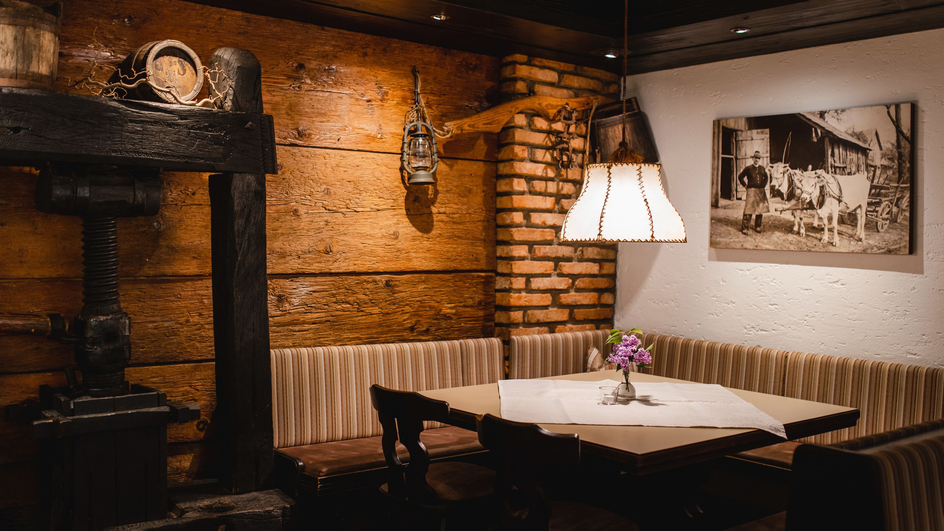Gemütliche Gaststube mit Holzwänden, einer Eckbank, einem Tisch mit Blumen und einer Lampe. An der Wand hängt ein historisches Foto.