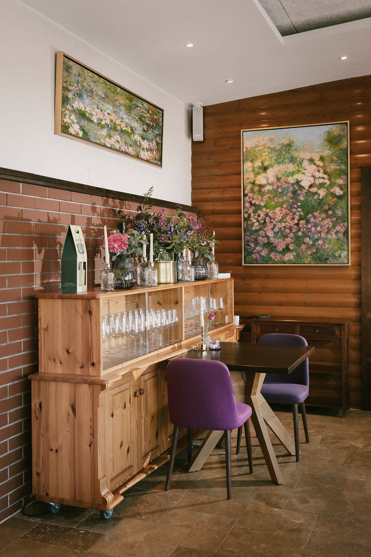 Ein Holzmöbel mit Gläsern, Blumenvasen und Kerzen steht an einer Wand. Daneben befinden sich ein Tisch mit zwei Stühlen sowie gerahmte Bilder an den Wänden.