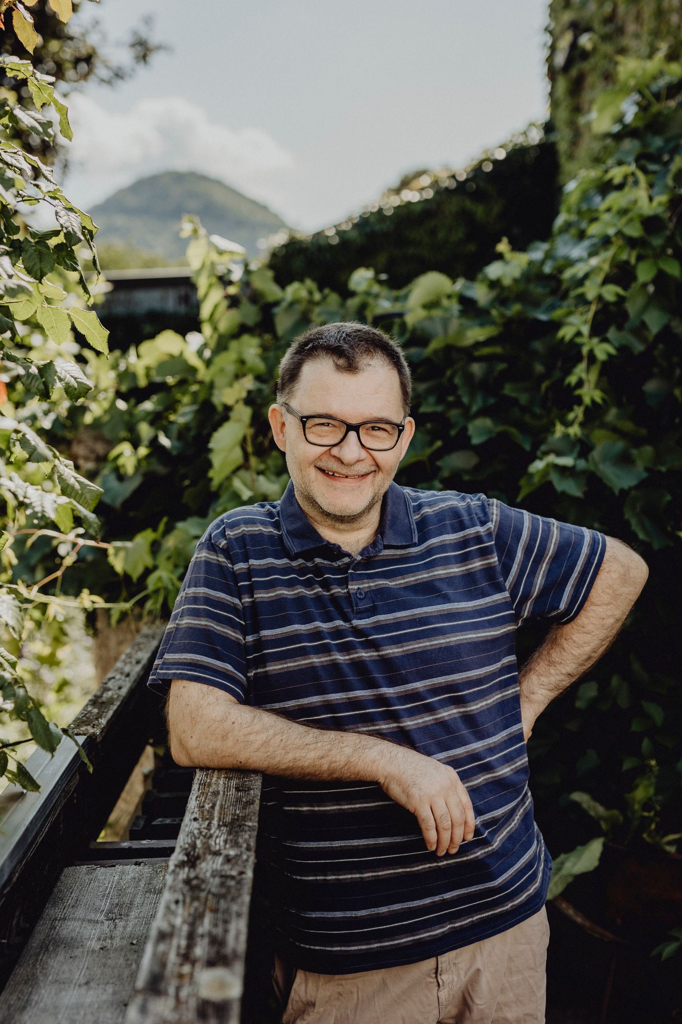 A man with glasses and a striped polo shirt stands smiling by a wooden fence surrounded by green plants.