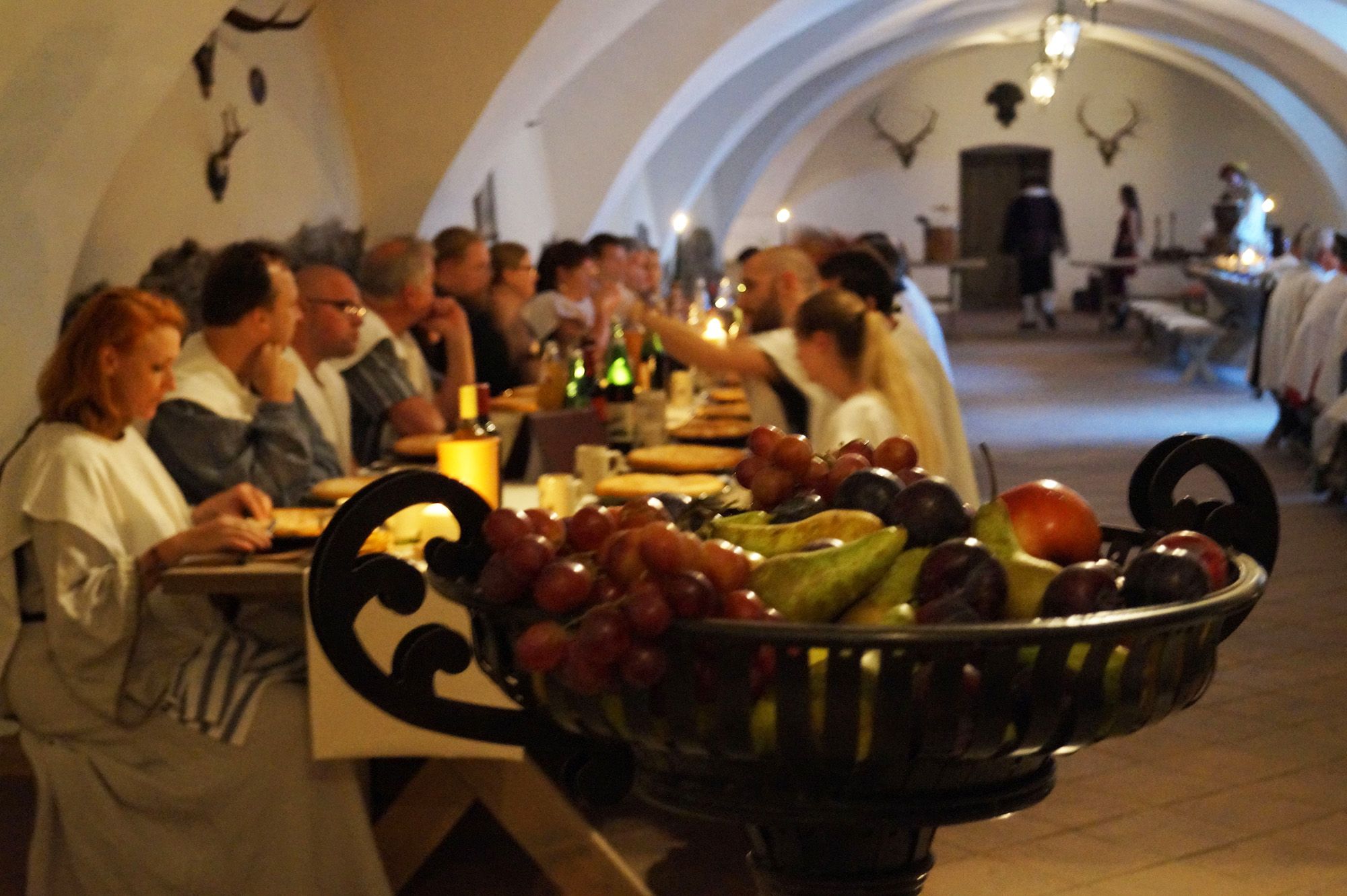 Menschen bei einem Ritteressen in einem Gewölbesaal mit Früchteschale im Vordergrund.