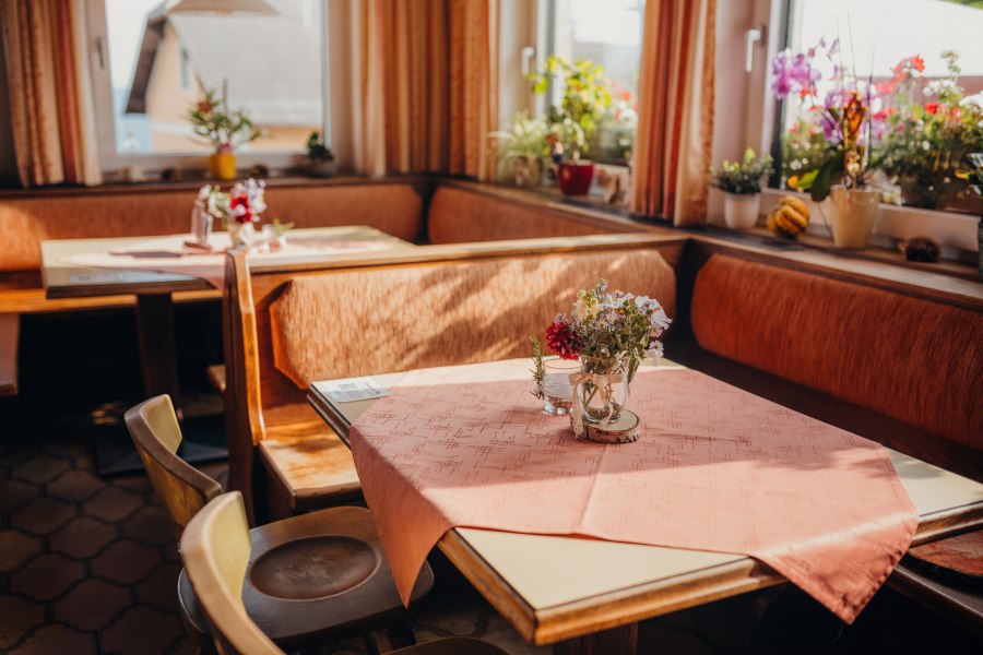 Gem&uuml;tliche Stube mit Holzm&ouml;beln, Tischdecken und Blumen auf den Tischen, umgeben von Fenstern mit Pflanzen.