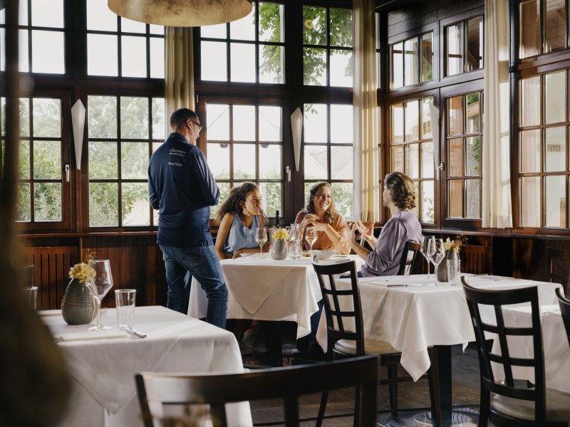 Innenansicht eines gemütlichen Restaurants mit großen Fenstern, drei Personen sitzen an einem Tisch und unterhalten sich, ein Kellner steht daneben.