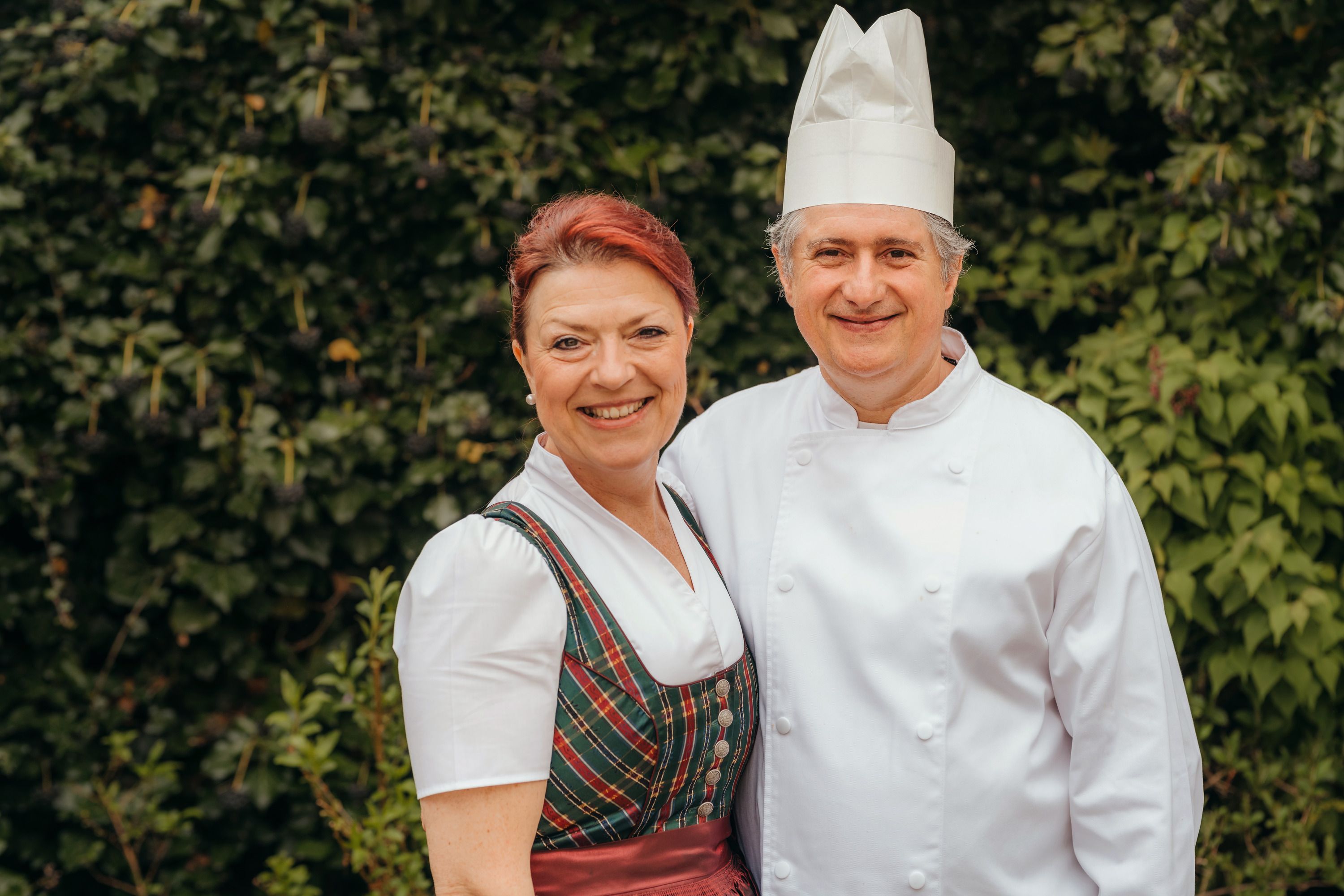Ein Mann in Kochkleidung und eine Frau in traditioneller Tracht lächeln in die Kamera, vor einem Hintergrund aus grünen Blättern.