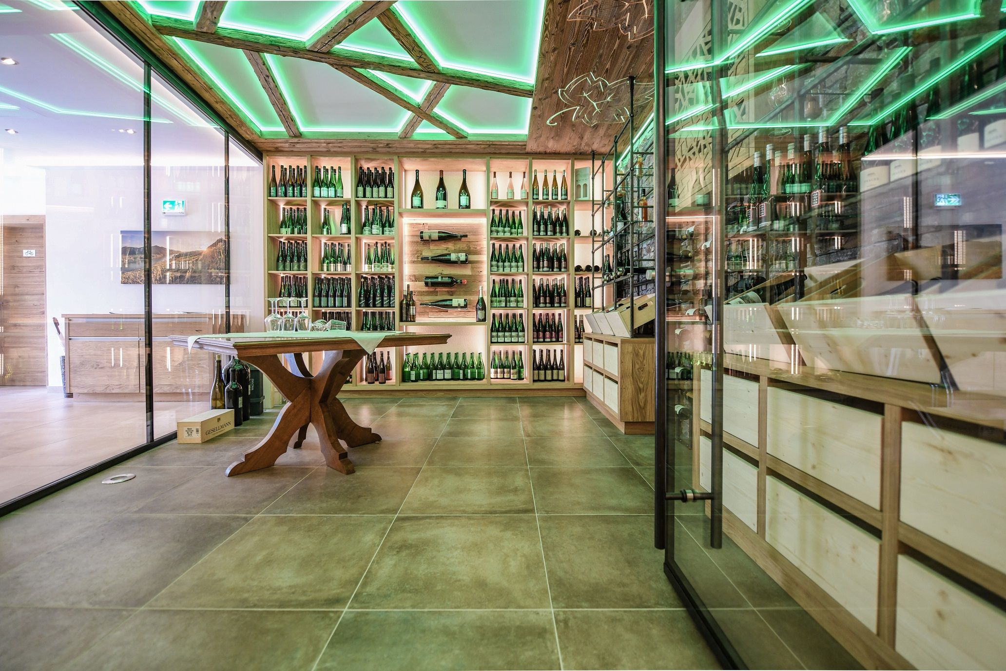 Moderner Weinpräsentationsraum mit Glaswänden, Holzregalen voller Weinflaschen und grün beleuchteter Decke.