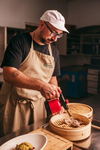 Ein Mann mit Sch&uuml;rze und M&uuml;tze flambiert Fisch in einem Bambusd&auml;mpfer.