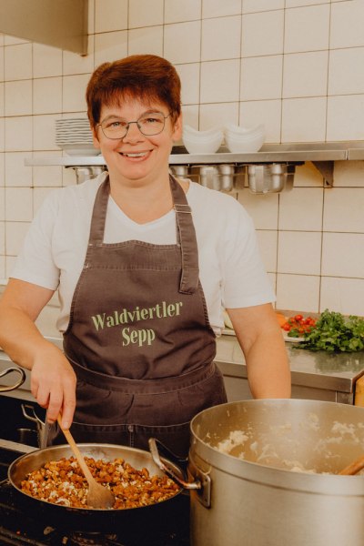 Eine K&ouml;chin in einer K&uuml;che mit Sch&uuml;rze, die "Waldviertler Sepp" tr&auml;gt, r&uuml;hrt in einer Pfanne.