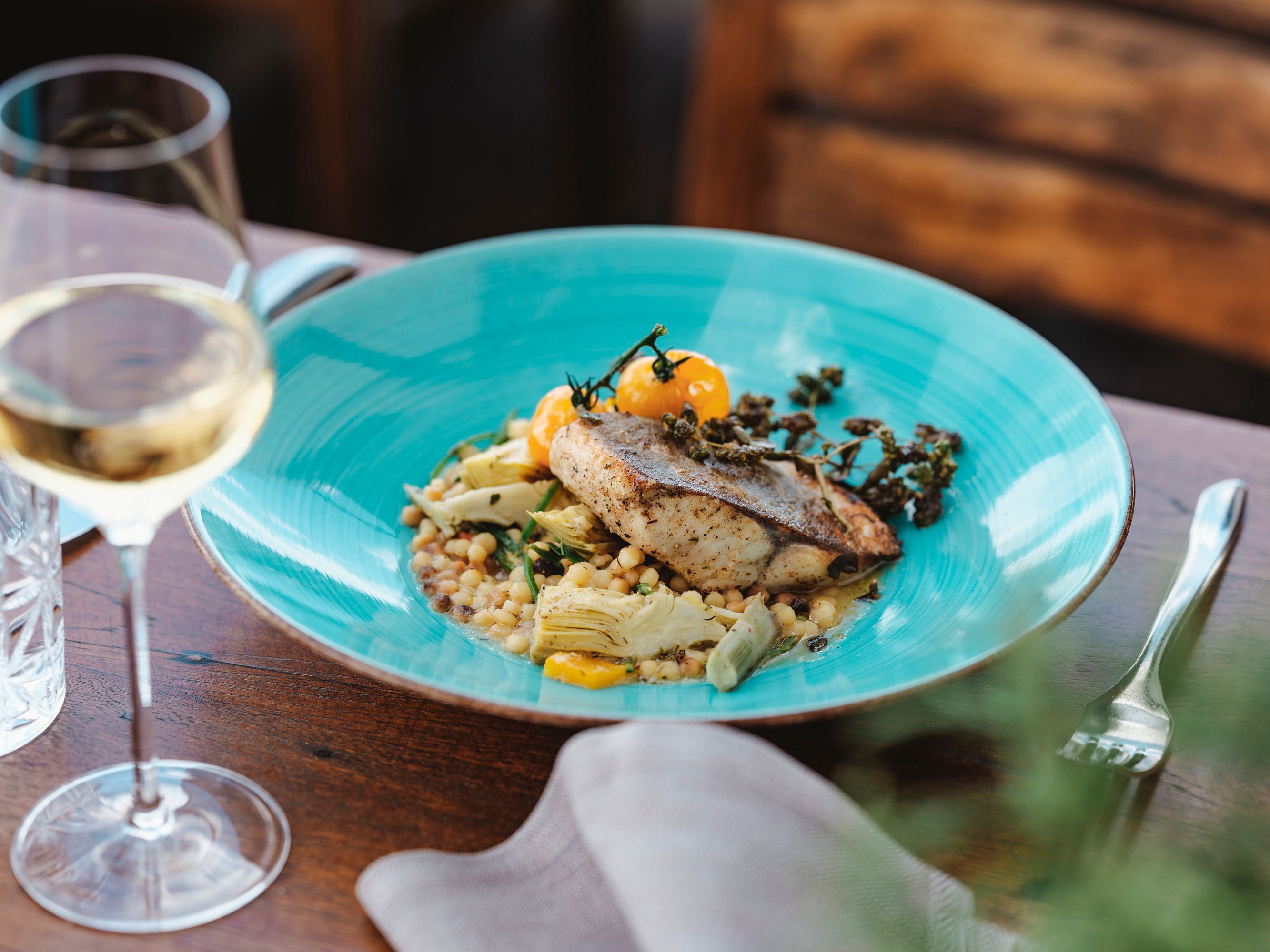 Elegantes Fischgericht mit Gemüse auf blauem Teller, Weißwein im Glas.