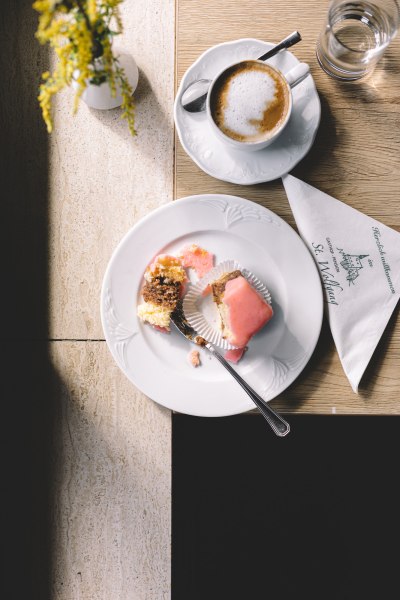 Ein Punschkrapferl auf einem Teller mit einer Gabel, daneben eine Tasse Kaffee und ein Glas Wasser auf einem Tisch.