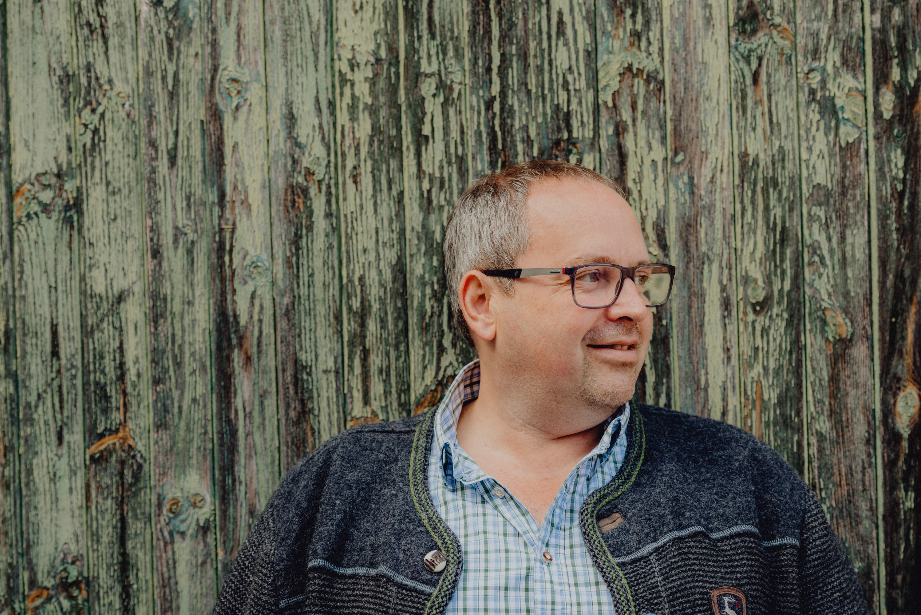 Mann mit Brille vor einer verwitterten Holzwand.
