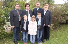 Eine Familie steht draußen in einem Garten, alle tragen formelle Kleidung. Im Hintergrund sind Büsche und Bäume zu sehen.