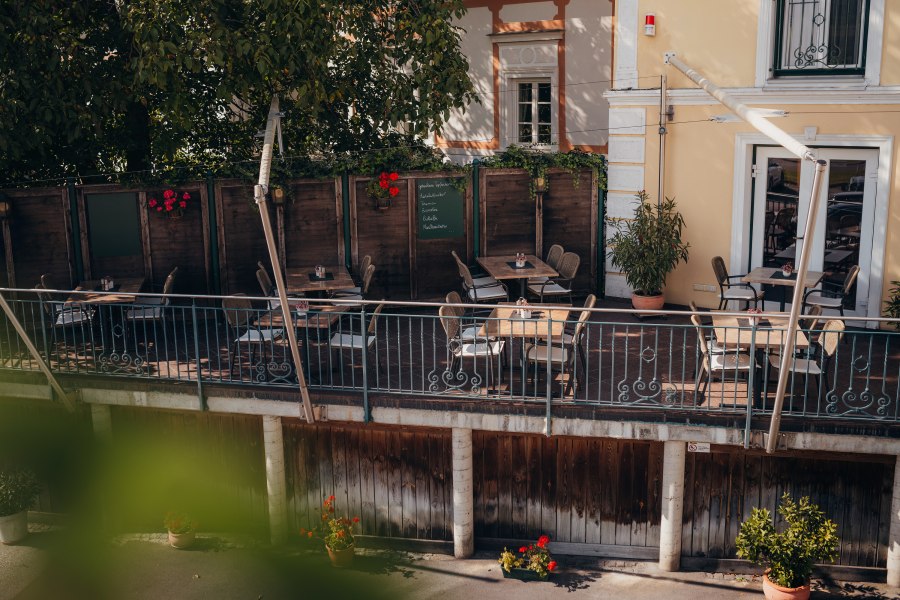 Gemütliche Terrasse eines Cafés mit Holztischen und Stühlen, umgeben von Pflanzen.