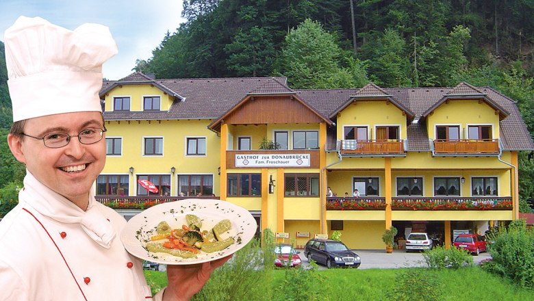 Ein Koch pr&auml;sentiert ein Gericht vor einem gelben Gasthof namens 'Zur Donaubr&uuml;cke'.