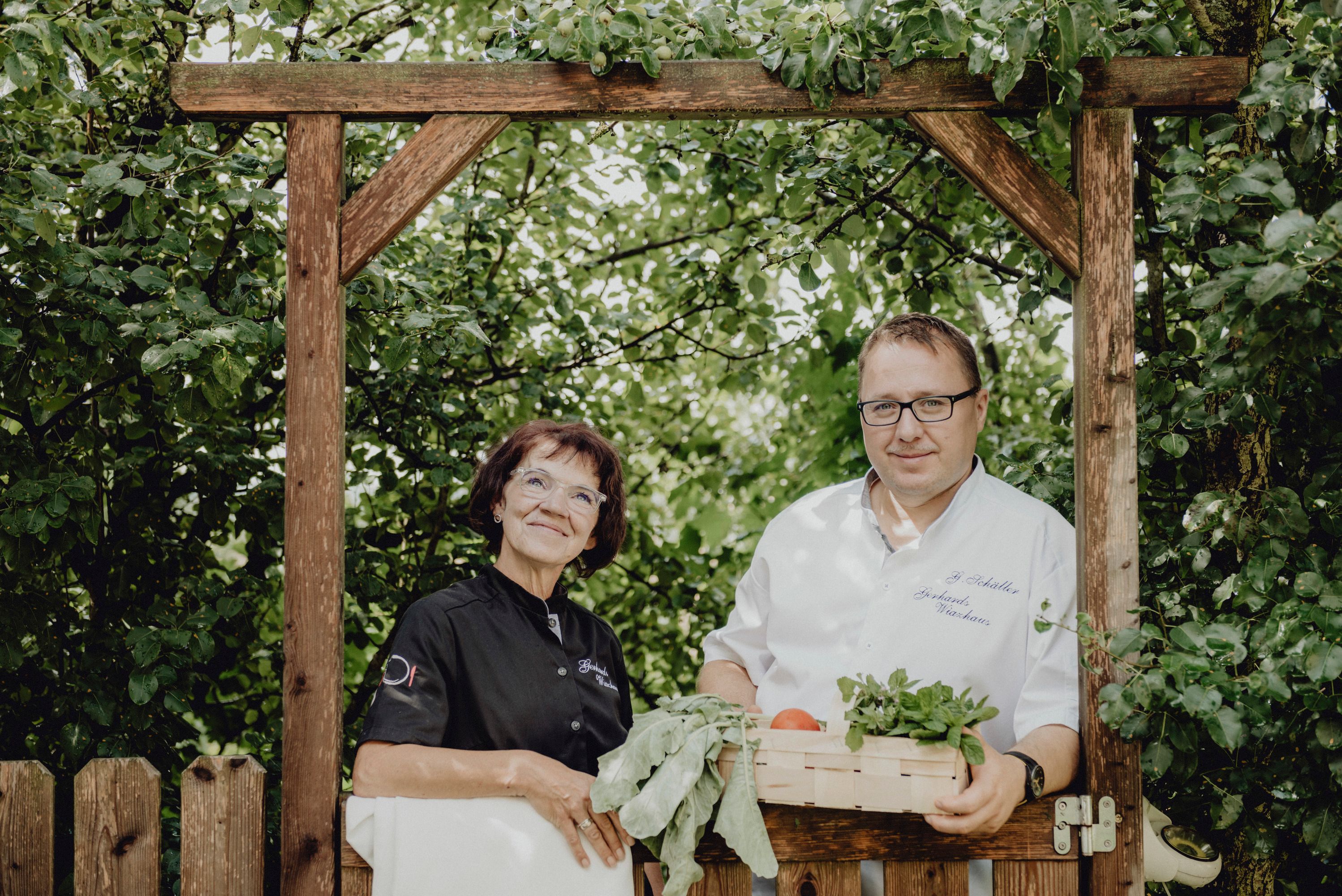 Zwei Personen stehen unter einem Holzbogen im Garten, eine Frau in schwarzer Kleidung und ein Mann in weißem Hemd mit einem Korb voller Gemüse.
