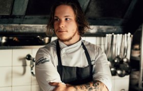 Junger Koch mit Tattoos in einer professionellen Küche, nachdenklich blickend.