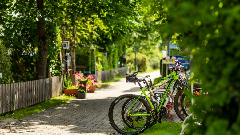 Fahrr&auml;der geparkt auf einem Radparkplatz bei einem Gasthof, umgeben von gr&uuml;nen B&auml;umen und Blumen.