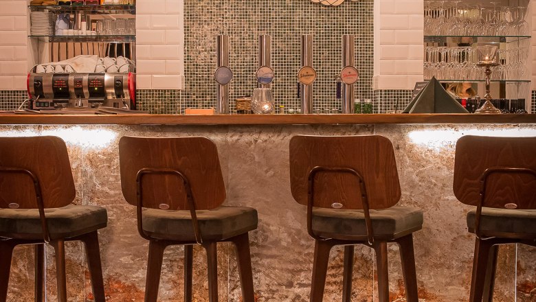 Innenansicht einer stilvollen Bar mit Holzst&uuml;hlen und beleuchtetem Tresen.