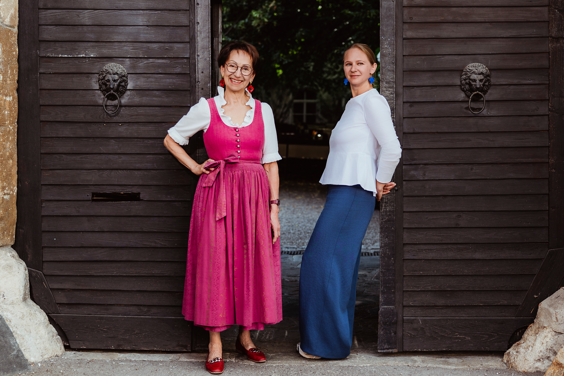 Zwei Personen stehen im Eingang eines Gebäudes mit großen dunklen Holztoren. Eine Person trägt ein pinkes Dirndl, die andere ein weißes Oberteil und einen langen blauen Rock.
