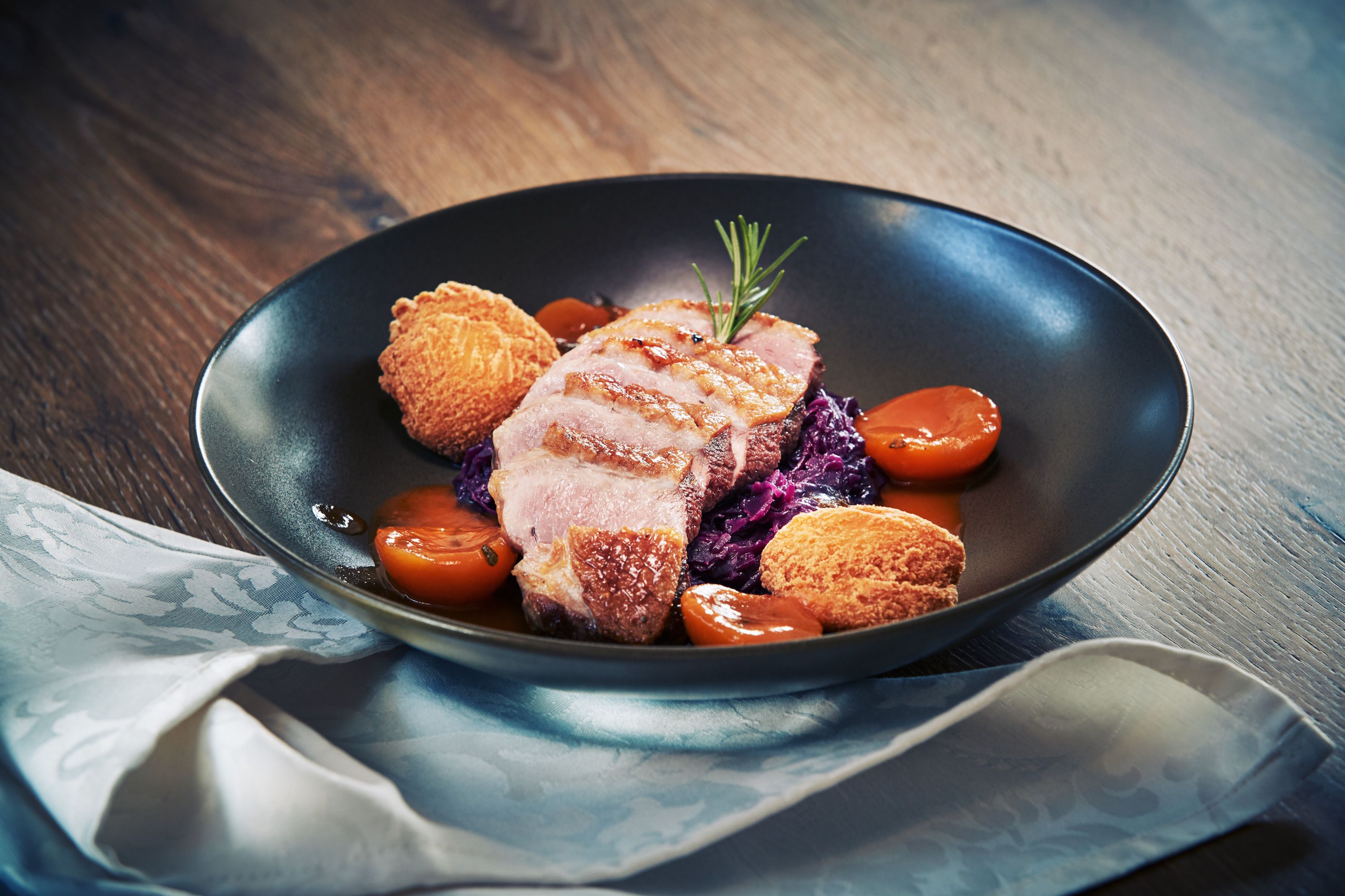 Gourmetgericht mit Entenbrust, Rotkraut, Kartoffelnocken und Aprikosen auf schwarzem Teller.
