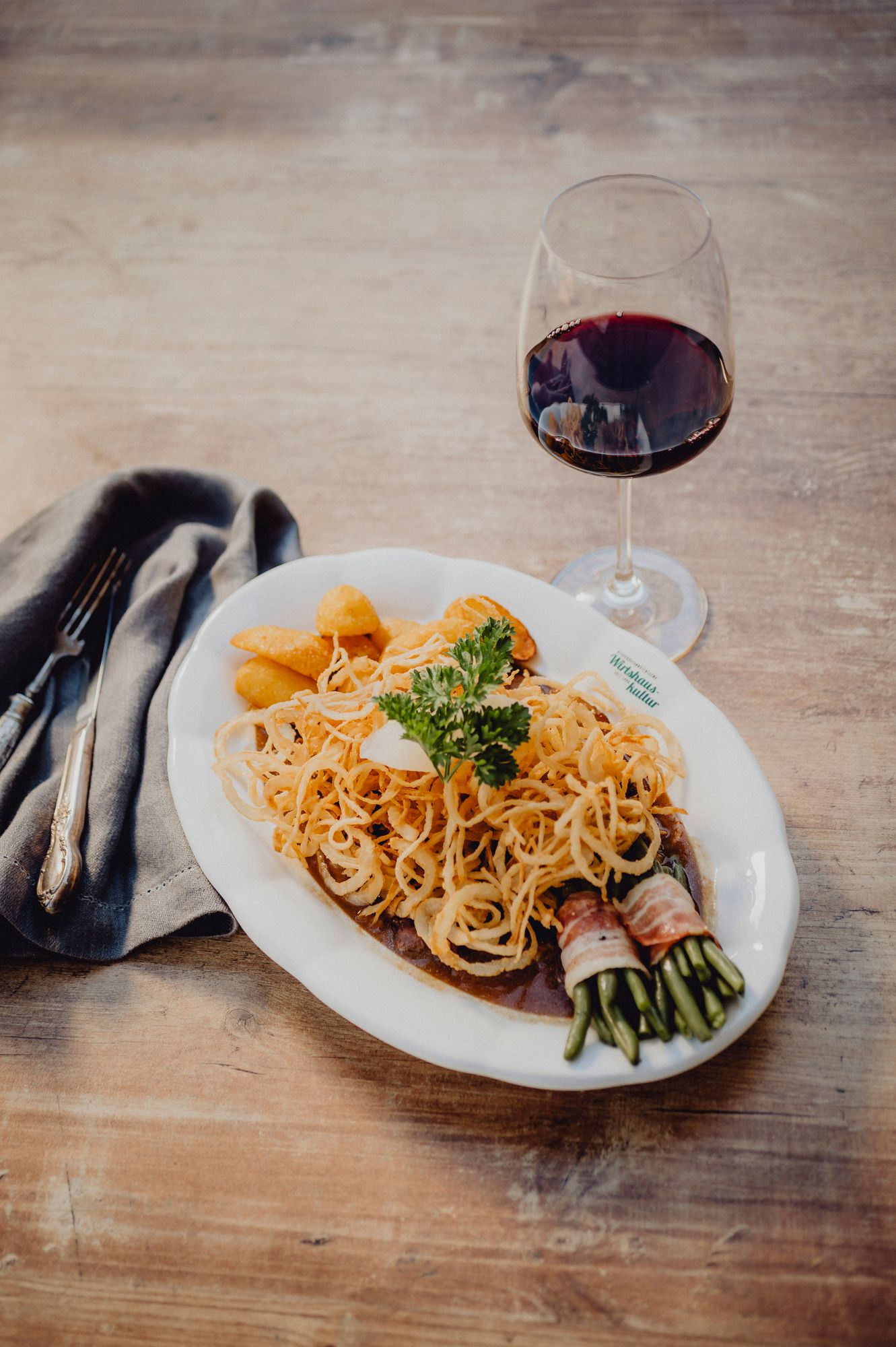 Ein Teller mit Zwiebelrostbraten, Kartoffeln und Speckbohnen, daneben ein Glas Rotwein auf einem Holztisch.