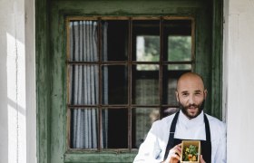 Ein Mann in Kochkleidung h&auml;lt eine Schachtel mit Kr&auml;utern vor einer gr&uuml;nen Holzt&uuml;r.