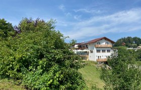 Ein wei&szlig;es Haus mit rotem Dach, umgeben von gr&uuml;nen B&auml;umen und Wiesen, unter blauem Himmel.