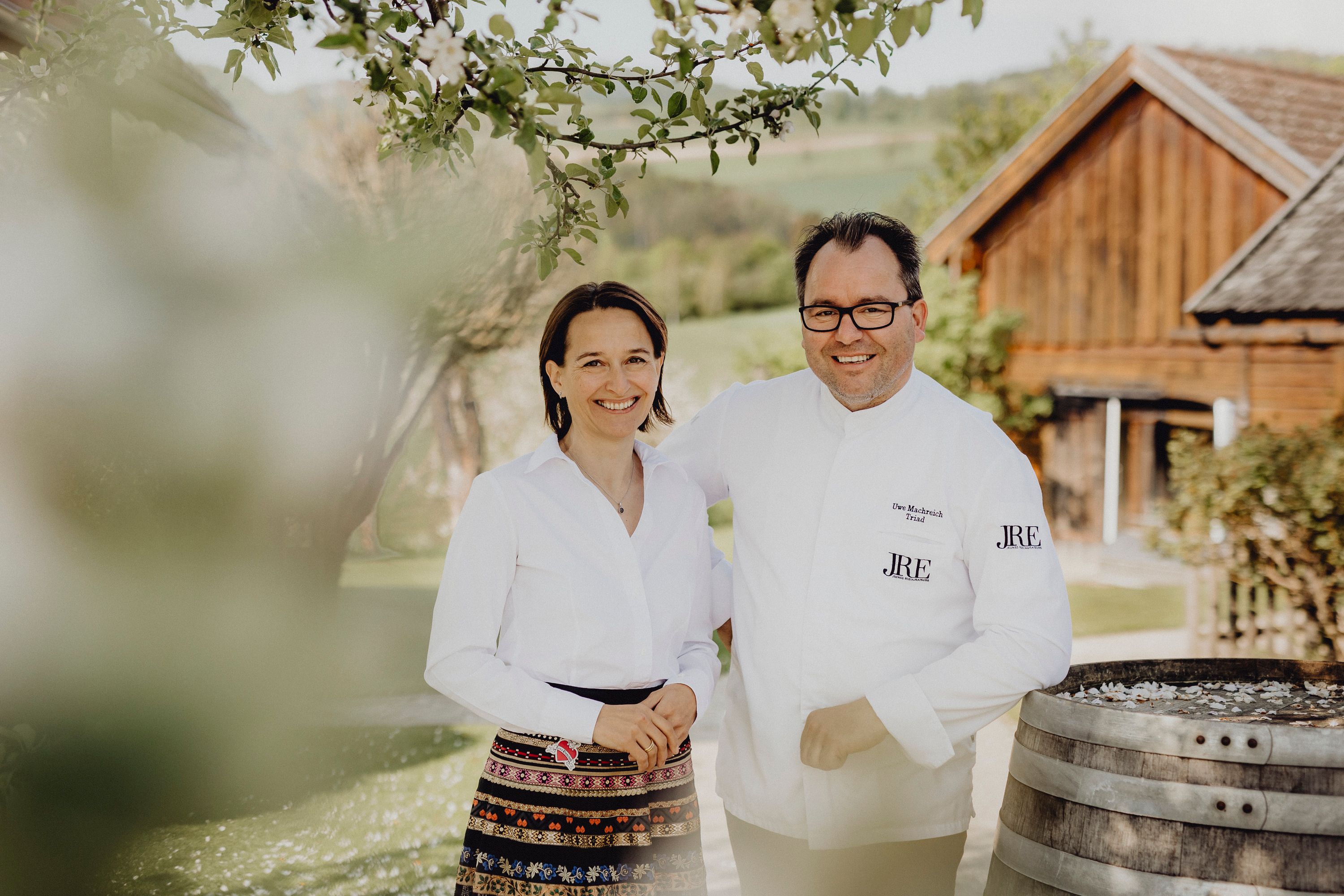 Ein Mann und eine Frau stehen lächelnd im Freien vor einem Holzgebäude.