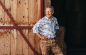 Mann in kariertem Hemd und Lederhose lehnt an Holzwand, lächelt.