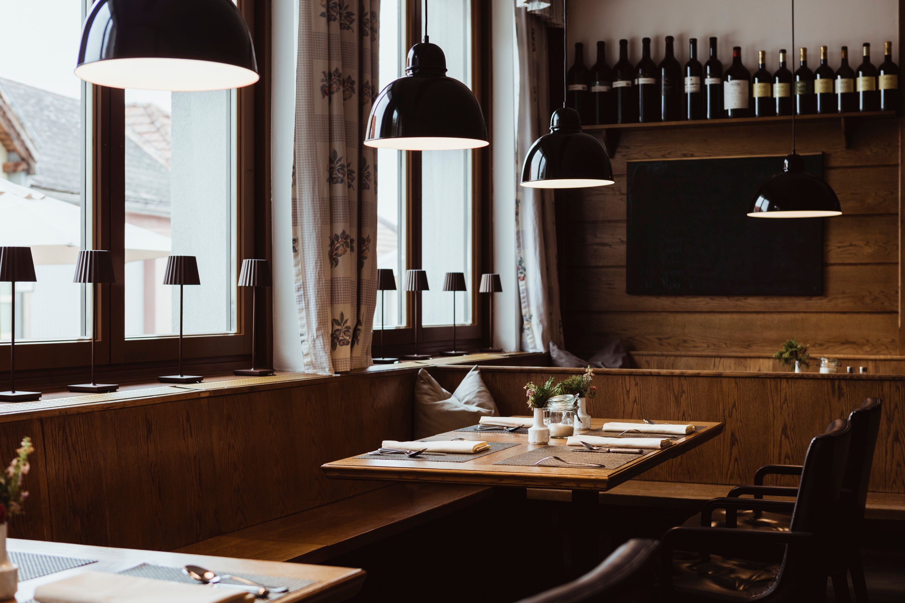 Gemütliches Restaurant mit Holztischen, Lampen und Weinflaschen an der Wand.