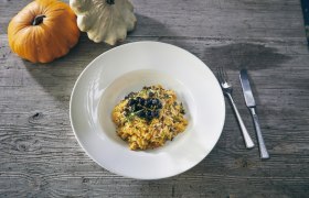 Ein Teller Kürbisrisotto mit Kräutern und Beeren, daneben zwei Kürbisse auf einem Holztisch.