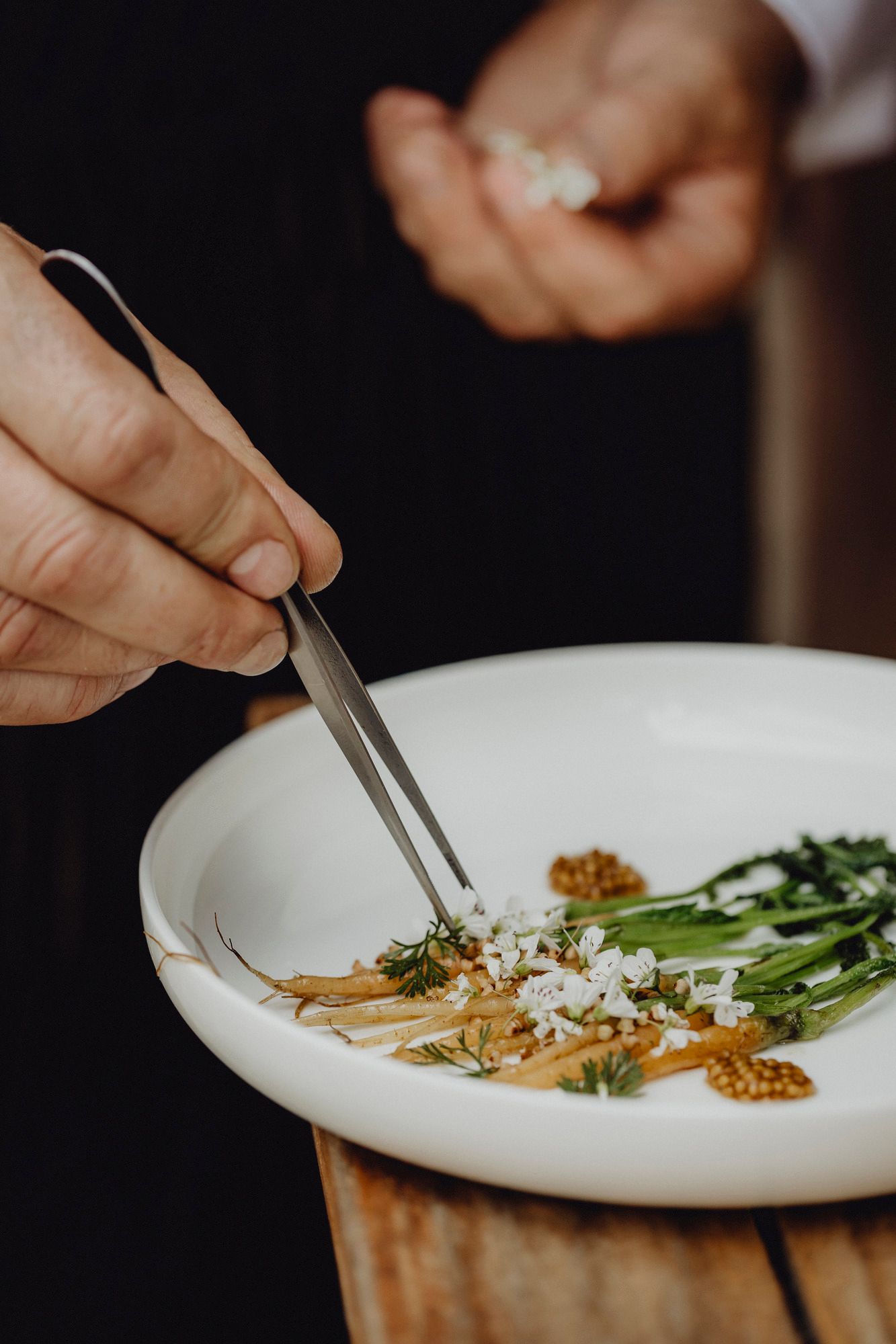 Nahaufnahme eines Tellers mit kunstvoll angerichteten Gemüsen und Blüten, eine Hand arrangiert mit einer Pinzette.