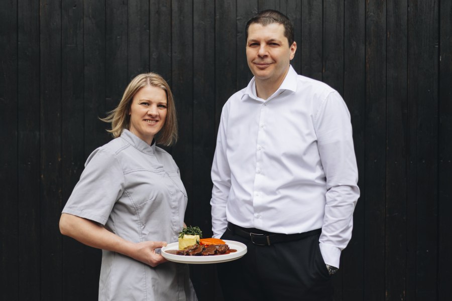 Zwei Personen, eine Frau in Kochkleidung mit einem Teller, vor dunklem Hintergrund.