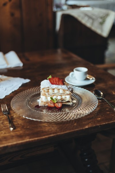 Glasteller mit Cremeschnitte, Erdbeeren und Minze, steht auf einem Holztisch neben einem Espresso.