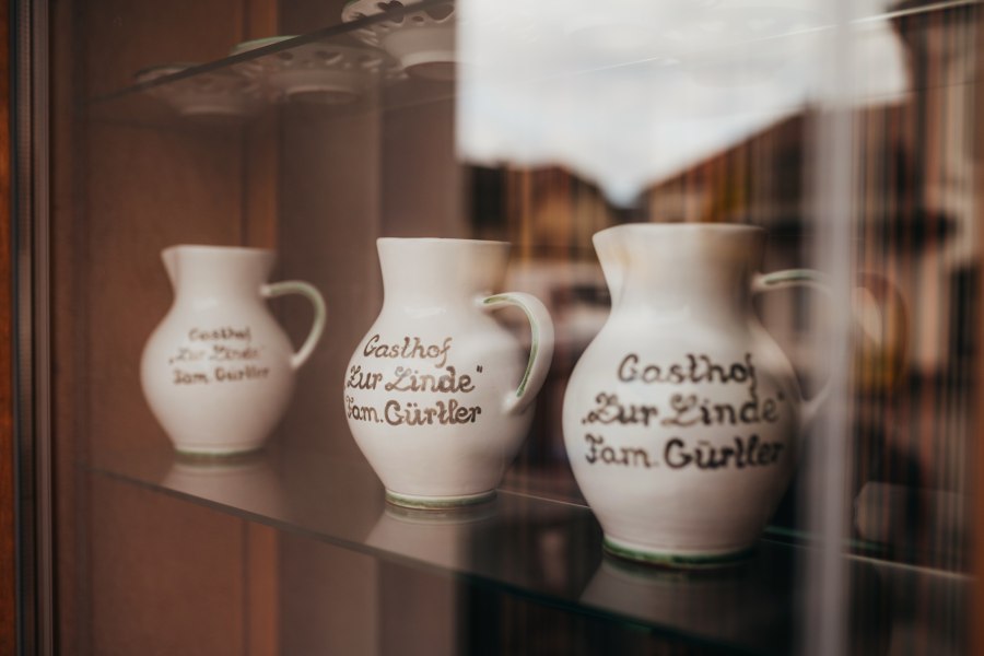 Drei Kr&uuml;ge mit Aufschrift "Gasthof Zur Linde Fam. G&uuml;rtler" in einer Vitrine.