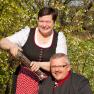 Ein Mann und eine Frau in traditioneller Kleidung lächeln im Freien, die Frau hält eine große Pfeffermühle.