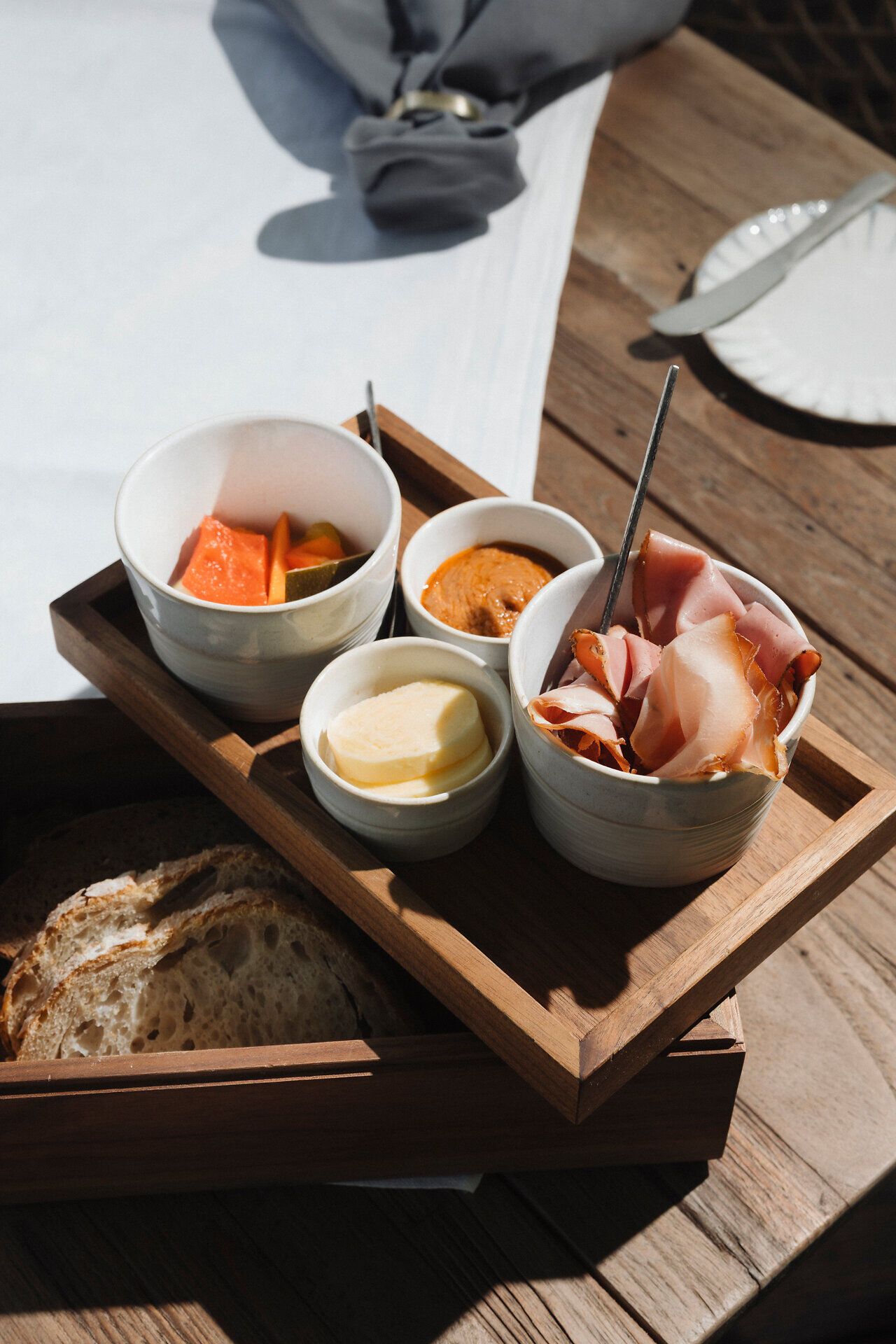 Ein Holztablett mit Brot sowie kleinen Schalen mit Aufstrichen, Butter, eingelegtem Gemüse und dünn geschnittenem Schinken steht auf einem Holztisch.
