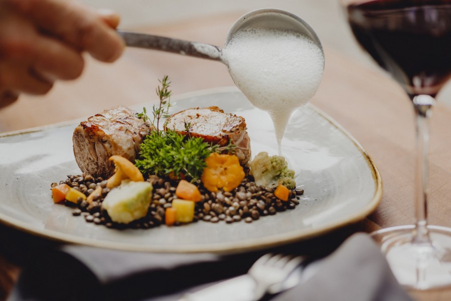 Schweinemedaillons mit Speckmantel, Linsen und Veltliner-Schaum auf einem Teller, daneben ein Glas Rotwein.