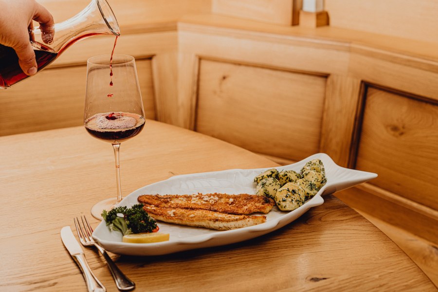 Ein Teller mit Zanderfilet und Kartoffeln in Knoblauchbutter, daneben ein Glas Rotwein auf einem Holztisch.