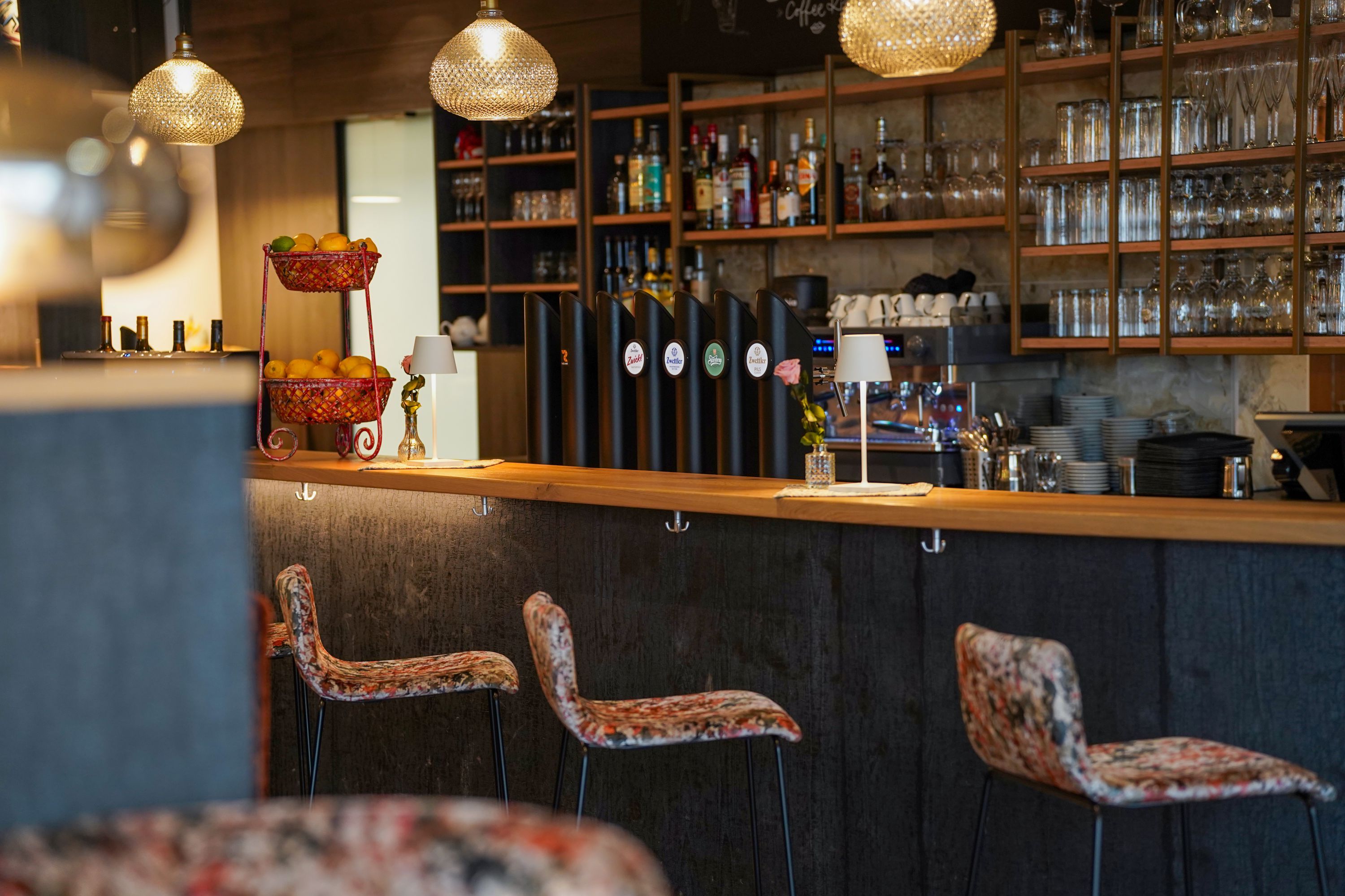 Gemütliche Bar im Innenraum mit Holzregalen, Gläsern, Flaschen, Pendelleuchten und Barhockern; auf dem Tresen steht ein Obstkorb.