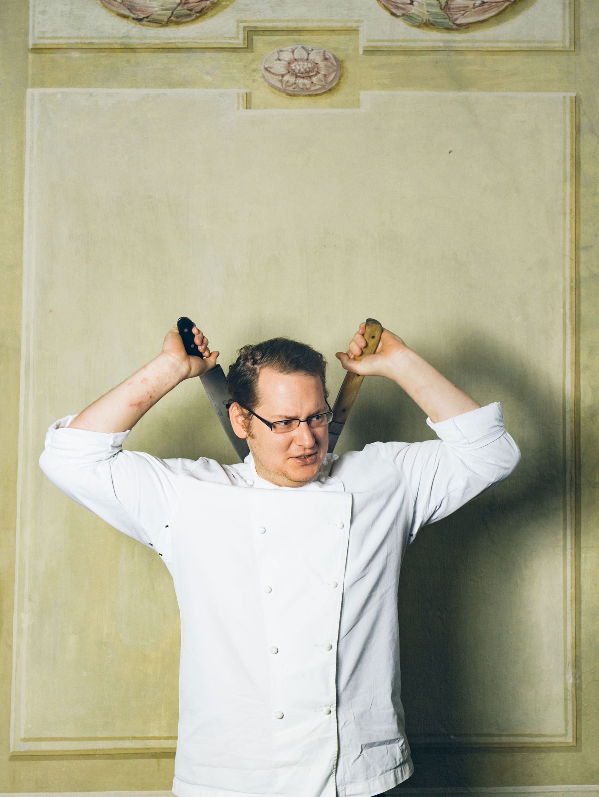 Ein Koch in weißer Uniform hält zwei Messer über dem Kopf.