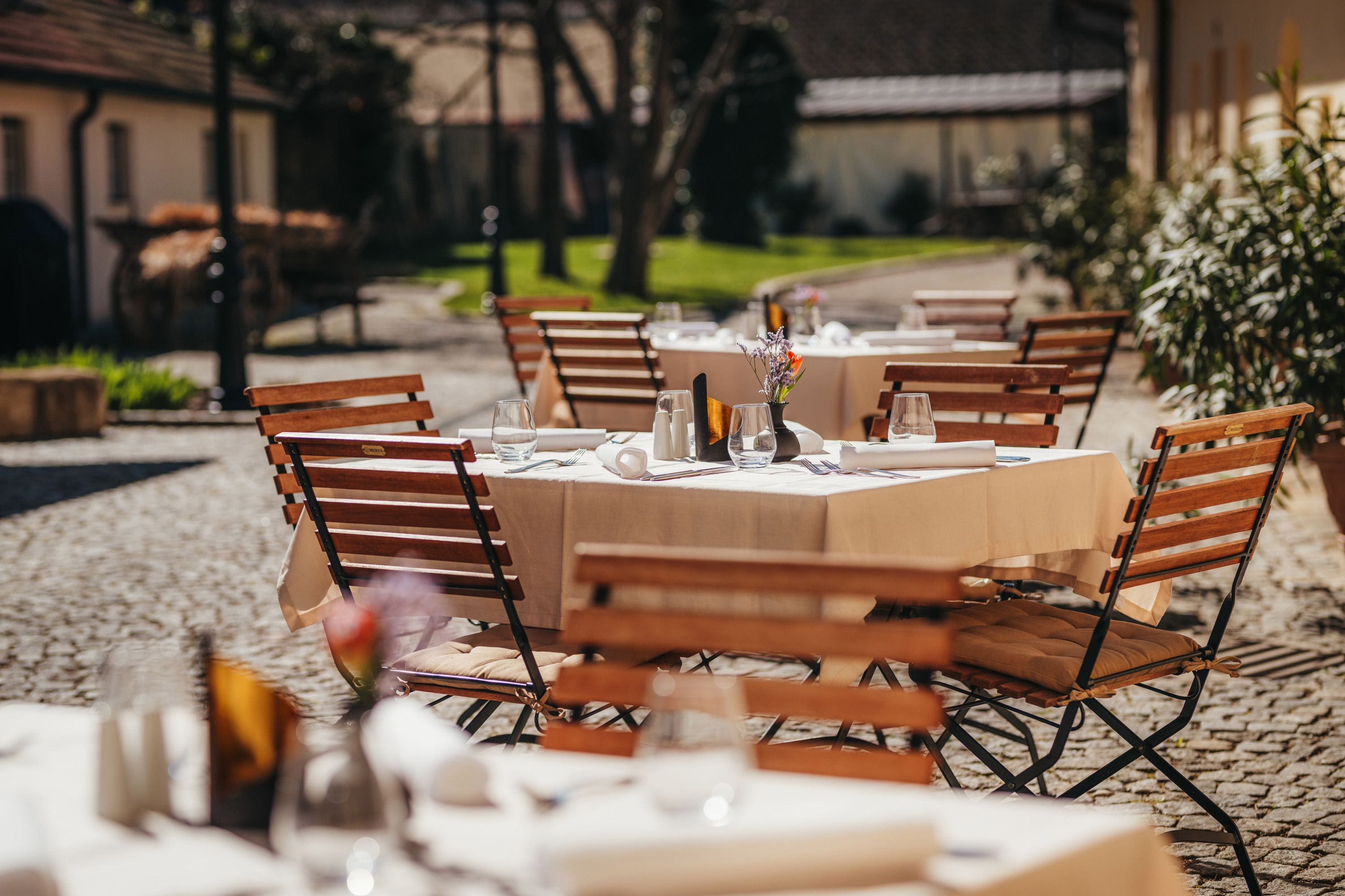 Ein sonniger Gastgarten mit Holztischen und -stühlen, gedeckt mit weißen Tischdecken und Gläsern, in einem gepflasterten Innenhof.