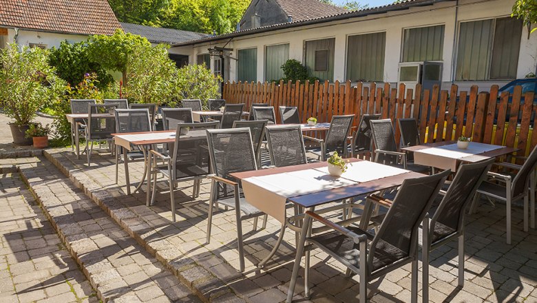 Gastgarten mit Tischen und St&uuml;hlen im Freien, umgeben von Pflanzen und einem Holzzaun.