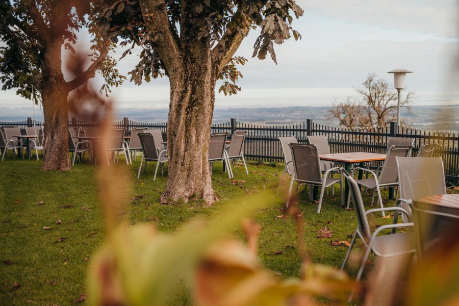 Garten mit Tischen und Stühlen unter Bäumen, mit Blick auf eine weite Landschaft.