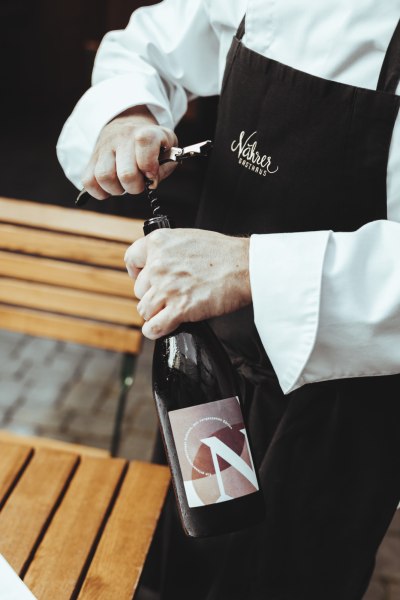 Person &ouml;ffnet eine Weinflasche mit einem Korkenzieher, tr&auml;gt eine Sch&uuml;rze mit der Aufschrift 'N&auml;hrer Gasthaus'.