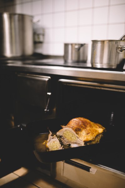 Ein Braten in einer schwarzen Ofenform wird aus einem Ofen gezogen. Im Hintergrund sind T&ouml;pfe auf einem Herd zu sehen.