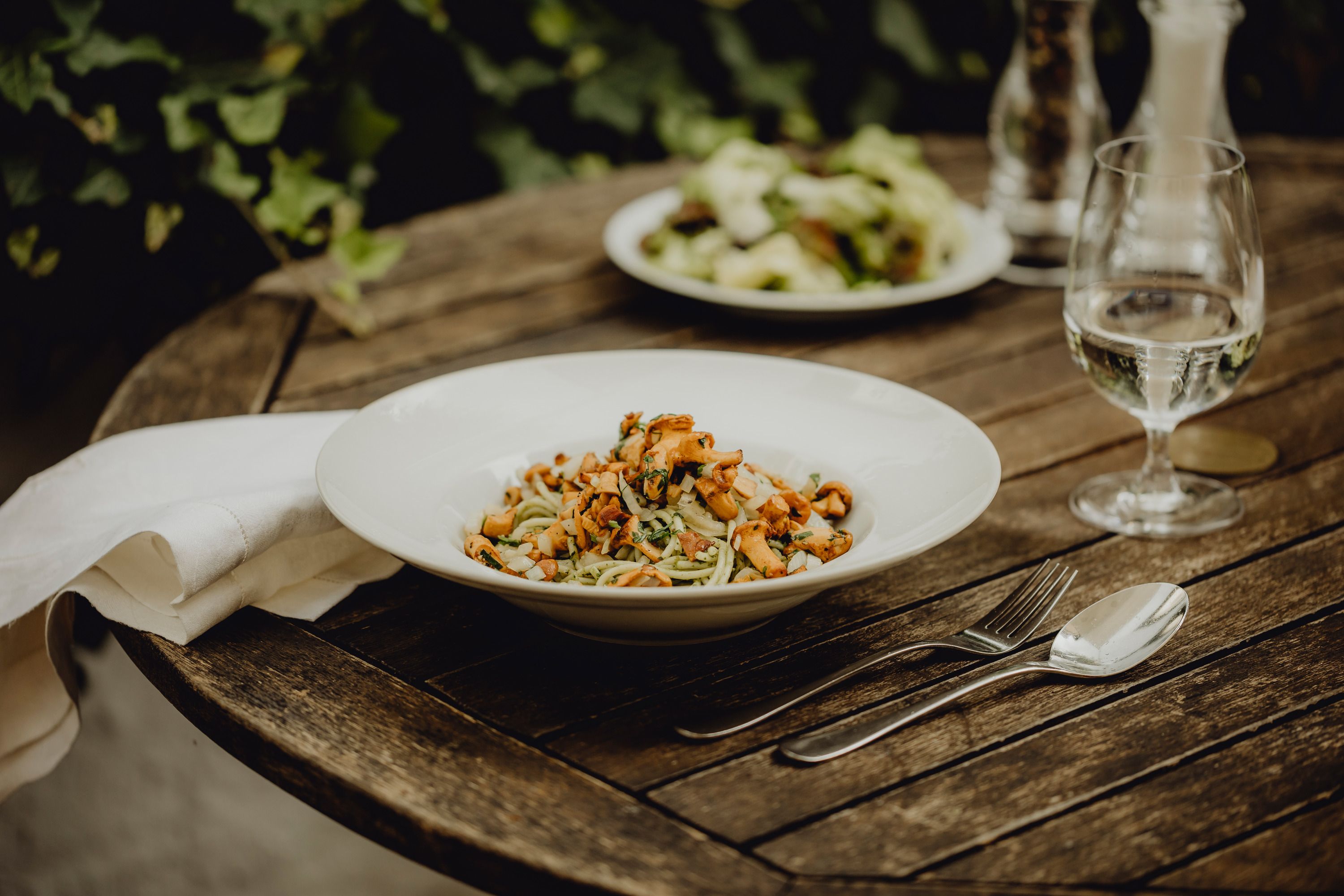 Ein Teller mit Pesto-Eierschwammerl-Nudeln auf einem Holztisch, daneben Besteck und ein Glas Wasser.