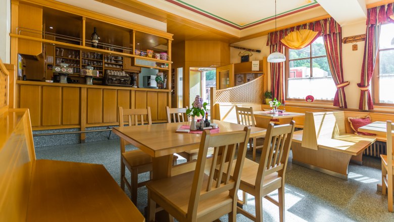 Gemütliches Gastzimmer mit Holzmöbeln, Tischen und Stühlen, dekoriert mit Blumen und Vorhängen, sonnendurchflutet durch große Fenster.