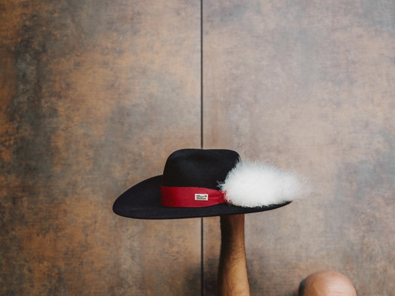Ein schwarzer Hut mit rotem Band und wei&szlig;er Feder wird von einer Hand hochgehalten.