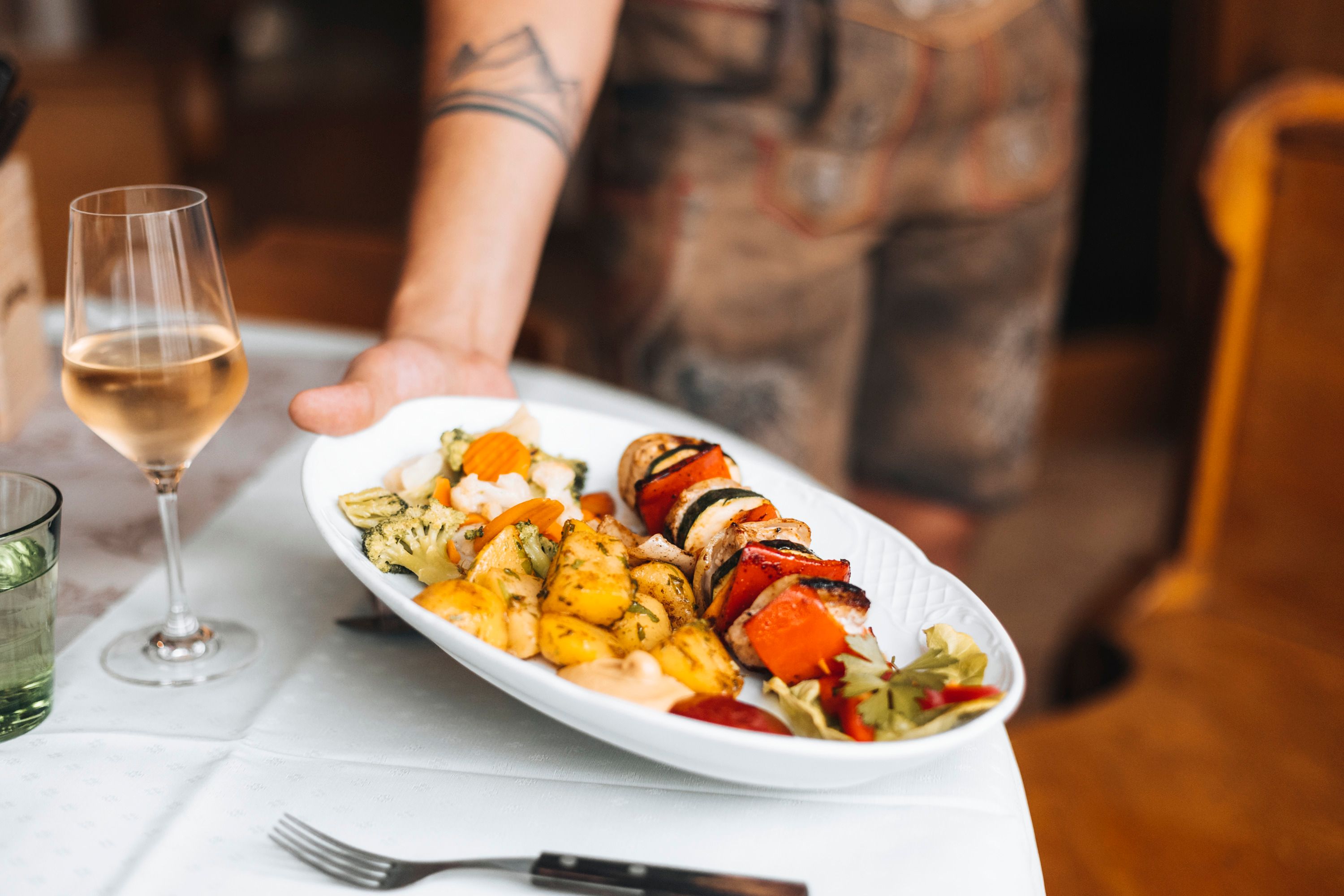 A plate of barbecue skewers, vegetables and potatoes is served, with a glass of white wine next to it.