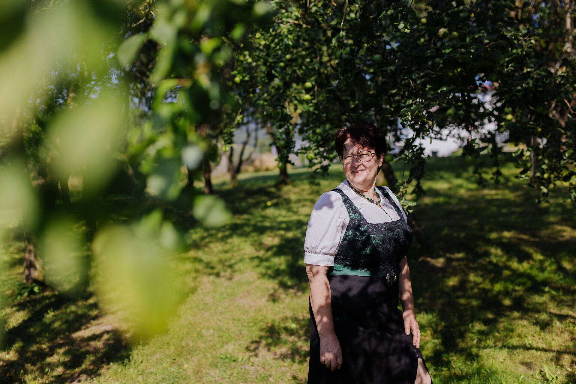 Inmitten der sanften Hügel und üppigen Wiesen strahlt die Sonne auf eine freundliche Dame, die in traditioneller Tracht lächelt. Die Bäume spenden Schatten und verleihen der Szenerie eine ruhige, einladende Atmosphäre, die zum Verweilen einlädt.
