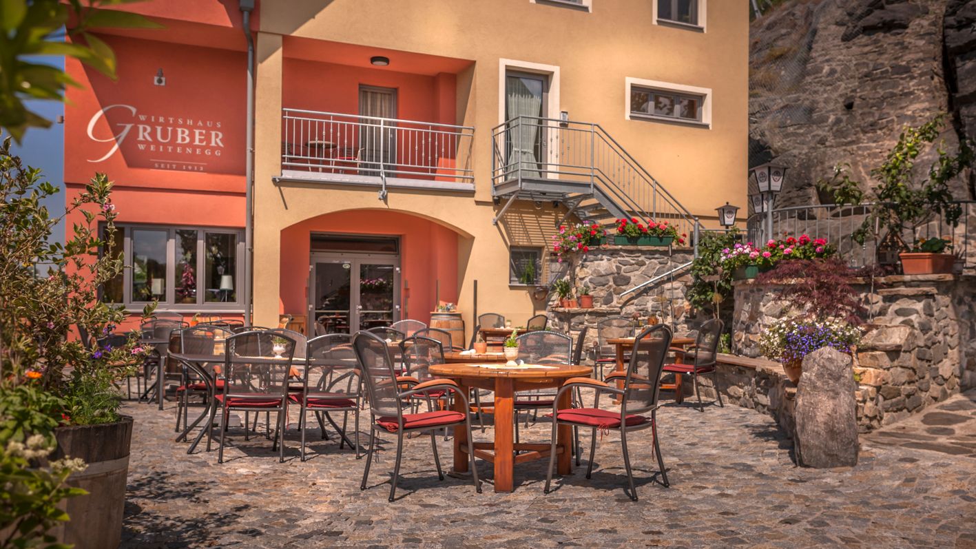 Außenbereich eines Restaurants mit Tischen und Stühlen auf einem gepflasterten Hof, umgeben von Blumen und Pflanzen.